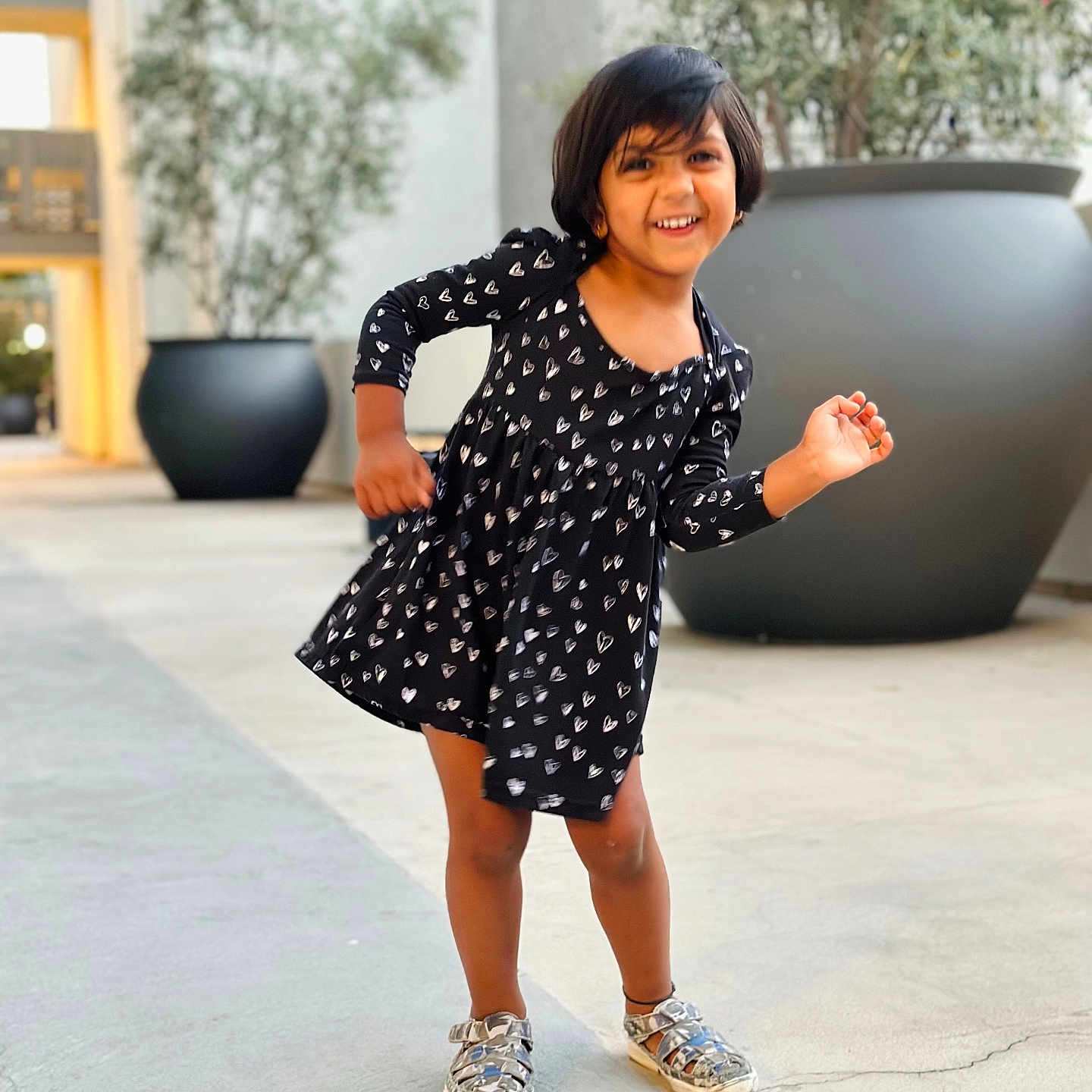 Ziona Yennam is registered to the contest to win money with this photo: background_blur, black_dress, casual_clothing, child, concrete, daylight, girl, happy, hearts_pattern, modern_building, outdoor, person, playful_pose, portrait, potted_plants, short_hair, silver_shoes, smiling, standing, urban