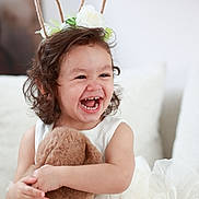 Leria participe au concours pour gagner de l'argent avec cette photo : child, toddler, laughing, curly_hair, bunny_ears, headband, teddy_bear, white_dress, couch, cushion, smile, happy, indoor, plush_toy, portrait, cute, soft_fabric, sitting, decor, person
