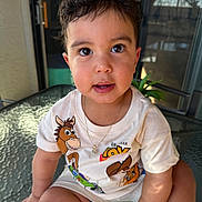 Neithan is registered to the contest to win money with this photo: balcony, big_eyes, bracelet, child, close_up, curly_hair, cute, glass_table, hand, happy, indoors_outdoors, necklace, plant, portrait, sitting, smiling, sunlit, toddler, toy_story_print, tshirt
