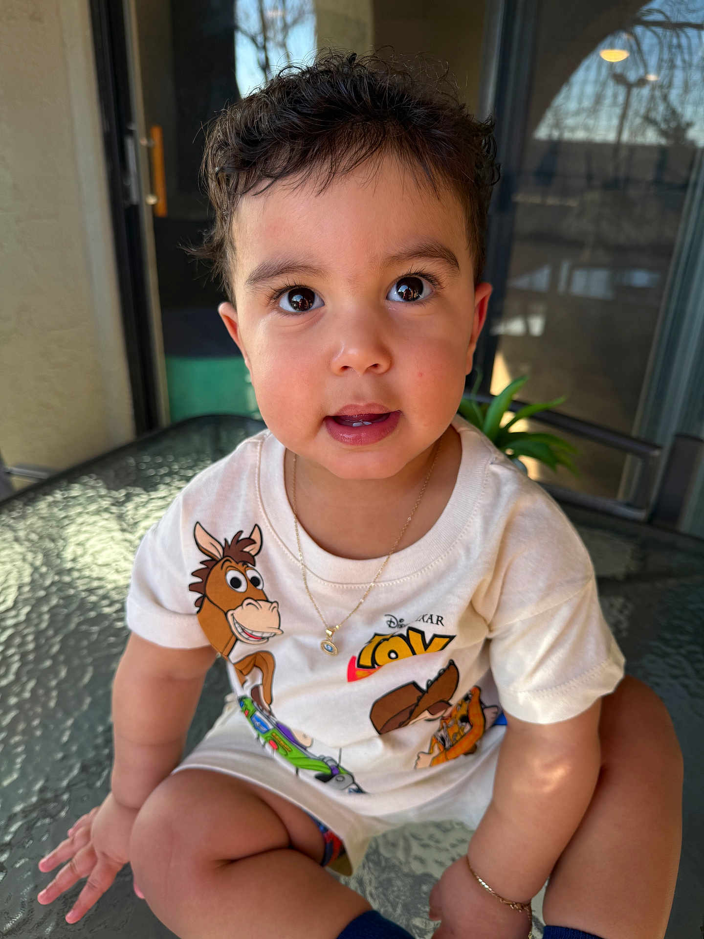 Neithan is registered to the contest to win money with this photo: balcony, big_eyes, bracelet, child, close_up, curly_hair, cute, glass_table, hand, happy, indoors_outdoors, necklace, plant, portrait, sitting, smiling, sunlit, toddler, toy_story_print, tshirt
