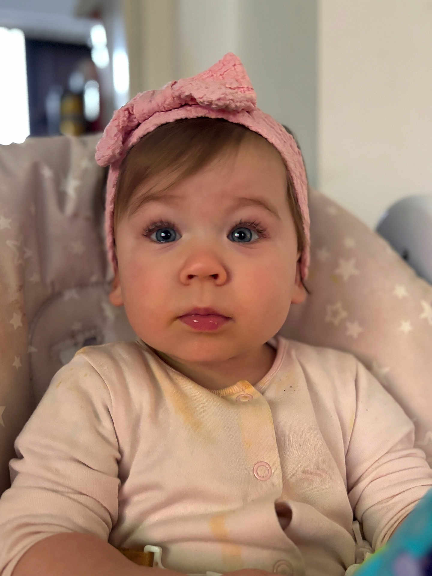 Freya is registered to the contest to win money with this photo: baby, child, pink_bow, headband, high_chair, onesie, face, blue_eyes, curious, seated, indoor, portrait, infant, cute, expression, soft_lighting, clothing, star_pattern, young_child, closeup