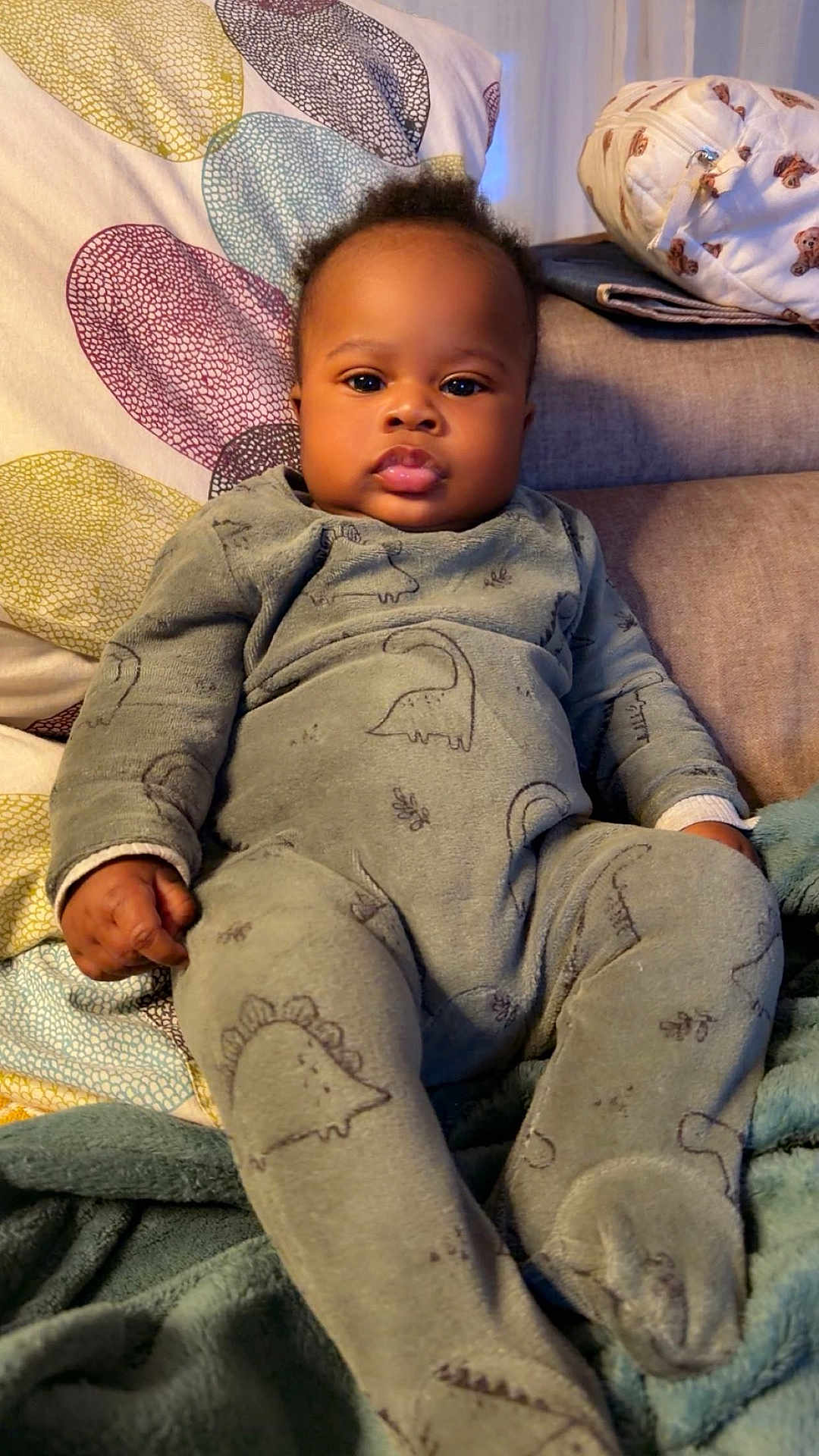 Joshua a rejoint le concours — aidez-le/la à gagner de superbes lots ! baby, infant, child, onesie, dinosaur_pattern, couch, blanket, pillow, indoors, cute, chubby_cheeks, relaxed, cozy, soft_fabric, portrait, young_child, seated, home, sleepwear, comfortable