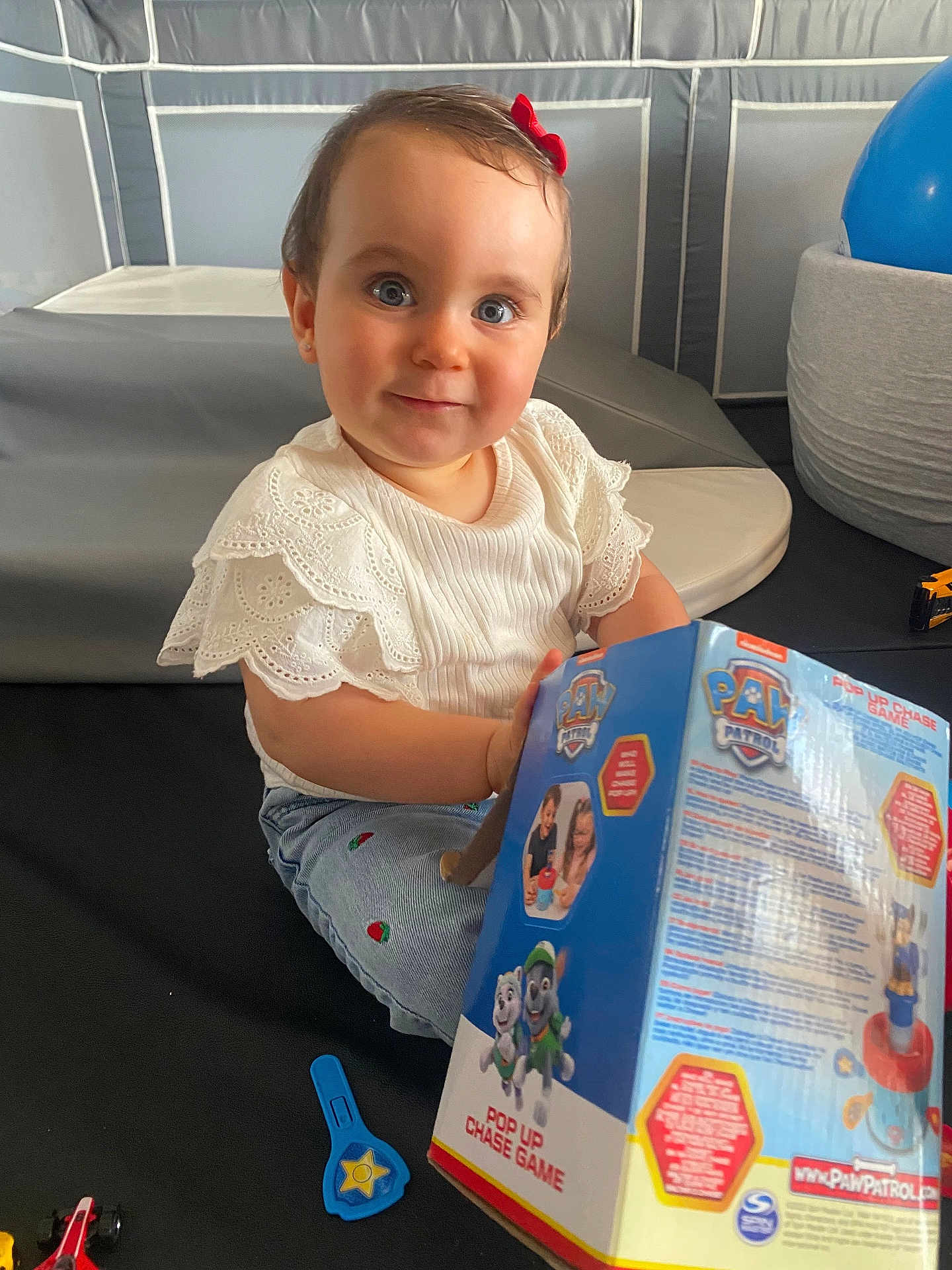 Charlize participe au concours pour gagner de l'argent avec cette photo : toddler, child, blue_eyes, red_bow, white_shirt, jeans, toy_box, paw_patrol, play_mat, indoor, smile, curious, cushion, toy, plastic_toy, childrens_toy, play_area, person, young_child, casual_clothing