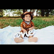 Axel is registered to the contest to win money with this photo: child, toddler, smiling, outdoor, grass, blanket, hat, birthday, one, sign, brown_clothing, bandana, shoes, nature, trees, cute, portrait, celebration, sitting, happy