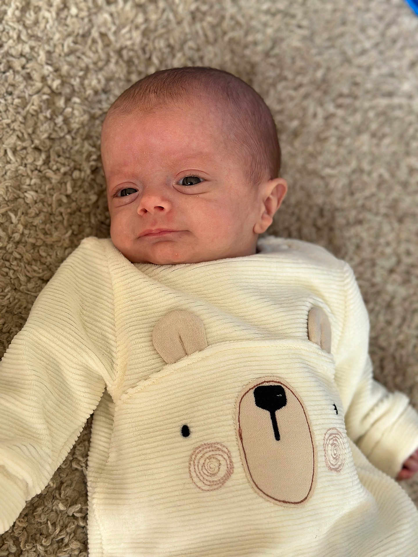 Mateo is registered to the contest to win money with this photo: baby, infant, person, face, clothing, onesie, bear_design, carpet, soft_texture, cute, newborn, child, portrait, indoors, laying_down, expression, apparel, warm, cozy, small