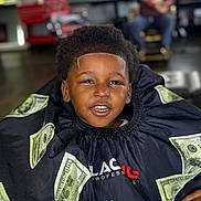 Kamari is registered to the contest to win money with this photo: child, boy, haircut, barber_cape, dollar_bills, smiling, portrait, barbershop, person, face, hair, clothing, happy, indoor, sitting, black_hair, young, casual, people, background