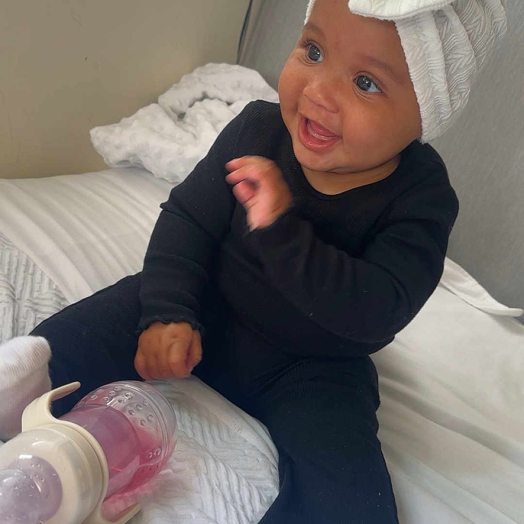 Reasiah Elohor is registered to the contest to win money with this photo: baby, bed, black_clothing, blanket, bottle, bow, child, cute, happy, headwrap, indoors, infant, natural_light, person, playful, portrait, smiling, toddler, white_socks, window