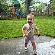 Camden-Rae joined the competition — help win amazing prizes! toddler, child, smiling, outdoor, grass, playground, slide, tree, fence, concrete, wet_surface, sandals, shorts, tshirt, stick, pigtails, person, daylight, nature, happy
