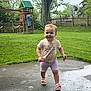 toddler, child, smiling, outdoor, grass, playground, slide, tree, fence, concrete, wet_surface, sandals, shorts, tshirt, stick, pigtails, person, daylight, nature, happy