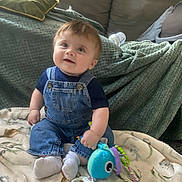 Edward joined the competition — help win amazing prizes! baby, child, infant, smiling, sitting, toy, plush_toy, blanket, denim_overalls, socks, couch, throw_blanket, bottle, indoor, cute, happy, person, face, young_child, playtime