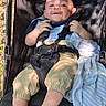 toddler, stroller, child, baby, smiling, outdoor, sunlight, shadow, blanket, clothing, feet, seatbelt, black, fabric, playful, relaxed, infant, person, portrait, sitting
