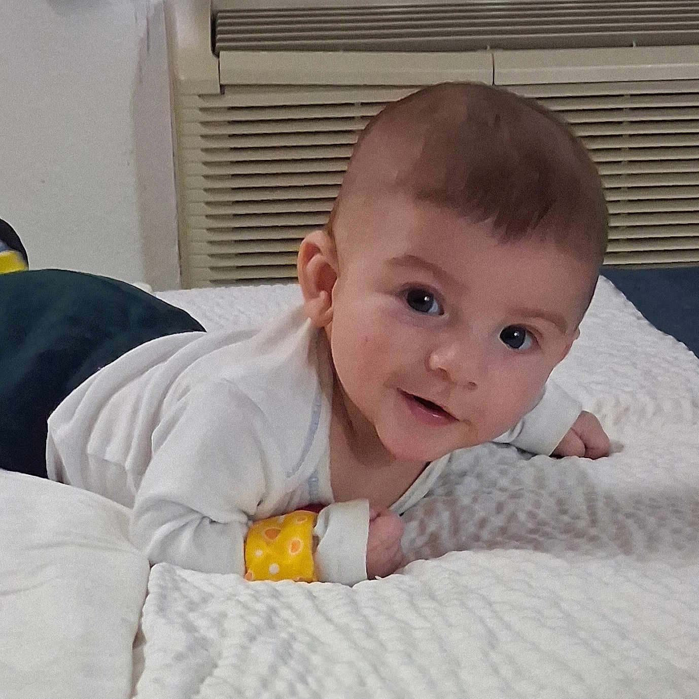 Jasper is registered to the contest to win money with this photo: baby, infant, child, blanket, white_clothing, dark_pants, colorful_socks, bracelet, smiling, lying_down, indoors, bedroom, carpet, face, head, eyes, hands, cute, portrait, young_child