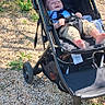 Jasper is registered to the contest to win money with this photo: toddler, stroller, outdoor, sunny, gravel, greenery, child, smiling, happy, casual_clothing, bare_feet, baby_seat, daylight, nature, leisure, relaxing, person, young_child, baby, transport