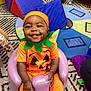 baby, carpet, child, costume, cute, festive, green_leaf_collar, happy, headwear, indoor, infant, orange_clothing, playful, playroom, pumpkin_costume, purple_chair, seated, small_person, smiling, toy