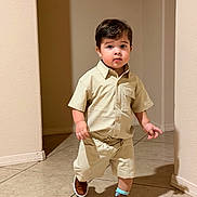 Gael is registered to the contest to win money with this photo: toddler, child, boy, walking, indoor, beige_clothing, shorts, shirt, brown_shoes, tile_floor, curious_expression, short_hair, casual_wear, home, person, portrait, young_child, cute, confident, neutral_background