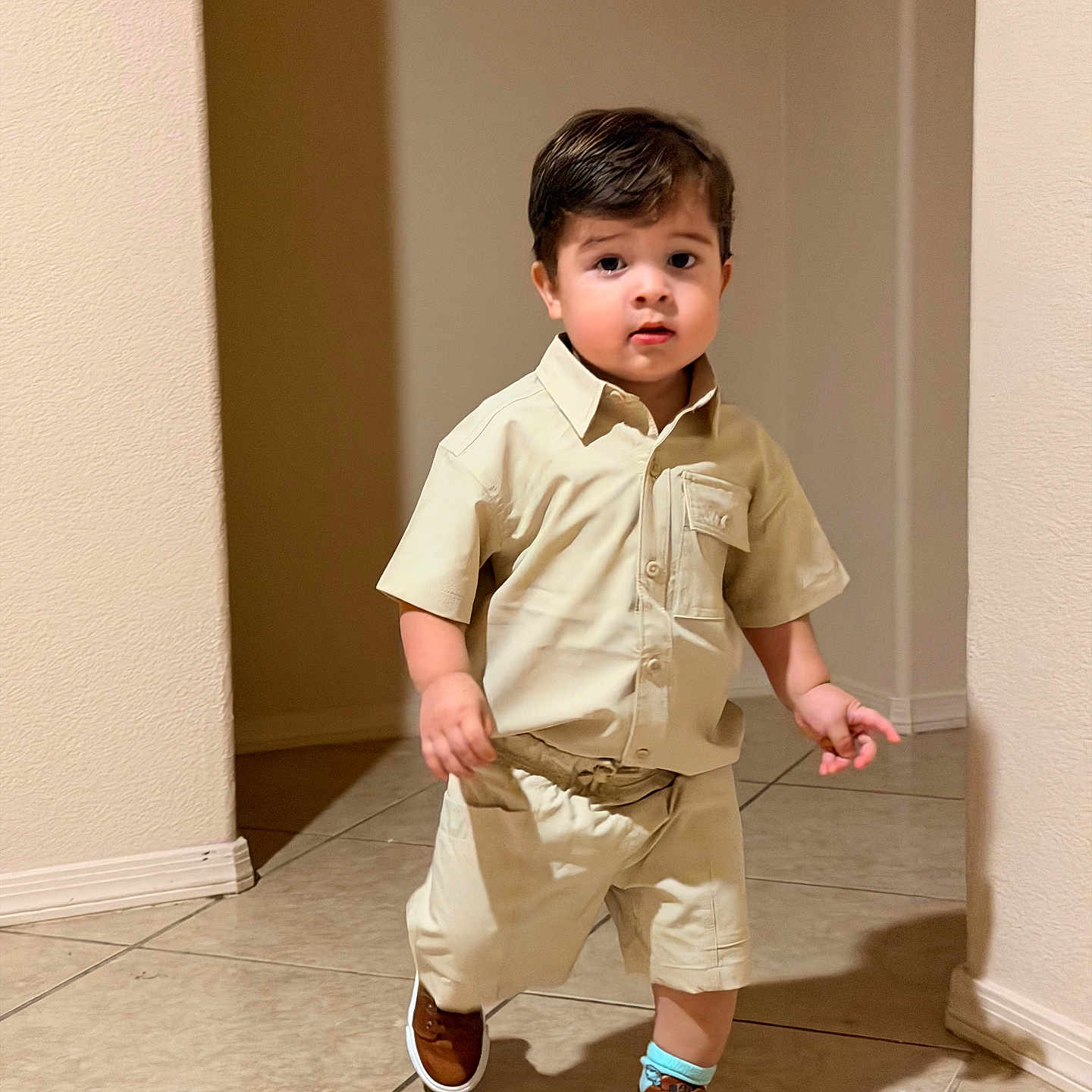 Gael is registered to the contest to win money with this photo: toddler, child, boy, walking, indoor, beige_clothing, shorts, shirt, brown_shoes, tile_floor, curious_expression, short_hair, casual_wear, home, person, portrait, young_child, cute, confident, neutral_background