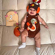 Winnie is registered to the contest to win money with this photo: baby, infant, couch, turkey_hat, brown_outfit, white_socks, wooden_sign, 2_months, pink_butterflies, squash, milestone, smiling, cute, indoor, soft_furniture, child, holiday_theme, portrait, celebration, cozy