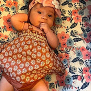 Aaliyah is registered to the contest to win money with this photo: baby, infant, child, headband, bow, dress, pattern, floral, mat, laying_down, cute, curious, hand, face, skin, legs, indoors, soft_lighting, portrait, person