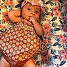 baby, infant, child, headband, bow, dress, pattern, floral, mat, laying_down, cute, curious, hand, face, skin, legs, indoors, soft_lighting, portrait, person