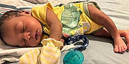 Amiri is registered to the contest to win money with this photo: baby, sleeping, bed, yellow_onesie, dinosaur_print, mittens, pacifier, blanket, pillow, infant, cute, peaceful, child, skin, foot, hand, indoors, rest, comfort, soft