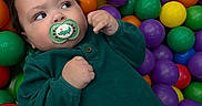 Branson is registered to the contest to win money with this photo: baby, child, pacifier, green_clothing, plastic_balls, colorful, play_area, indoor, cute, infant, face, hands, looking_away, purple_balls, yellow_balls, orange_balls, green_balls, red_balls, toy, playtime