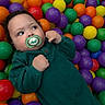 baby, child, pacifier, green_clothing, plastic_balls, colorful, play_area, indoor, cute, infant, face, hands, looking_away, purple_balls, yellow_balls, orange_balls, green_balls, red_balls, toy, playtime