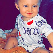 Maylon a rejoint le concours — aidez-le/la à gagner de superbes lots ! toddler, child, smiling, blue_eyes, white_clothing, print_text, blanket, orange_blanket, couch, indoor, sitting, happy, face, hair, person, cozy, cute, young_child, portrait, comfort