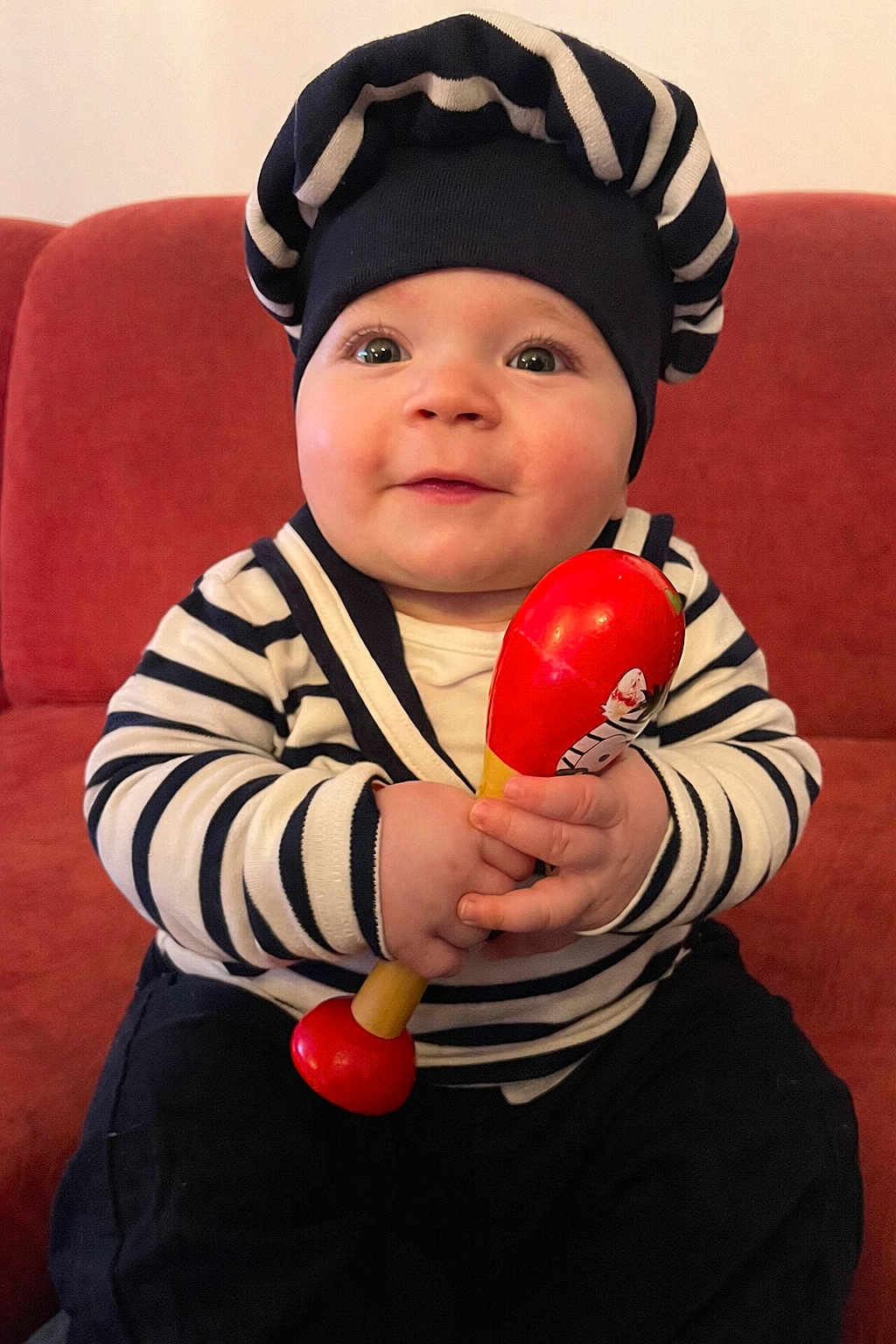 Alessio a rejoint le concours — aidez-le/la à gagner de superbes lots ! baby, infant, child, striped_clothing, striped_hat, navy_hat, red_toy, wooden_rattle, maraca, sofa, couch, sitting, portrait, smile, chubby_cheeks, big_eyes, hands, indoor, cute, outfit