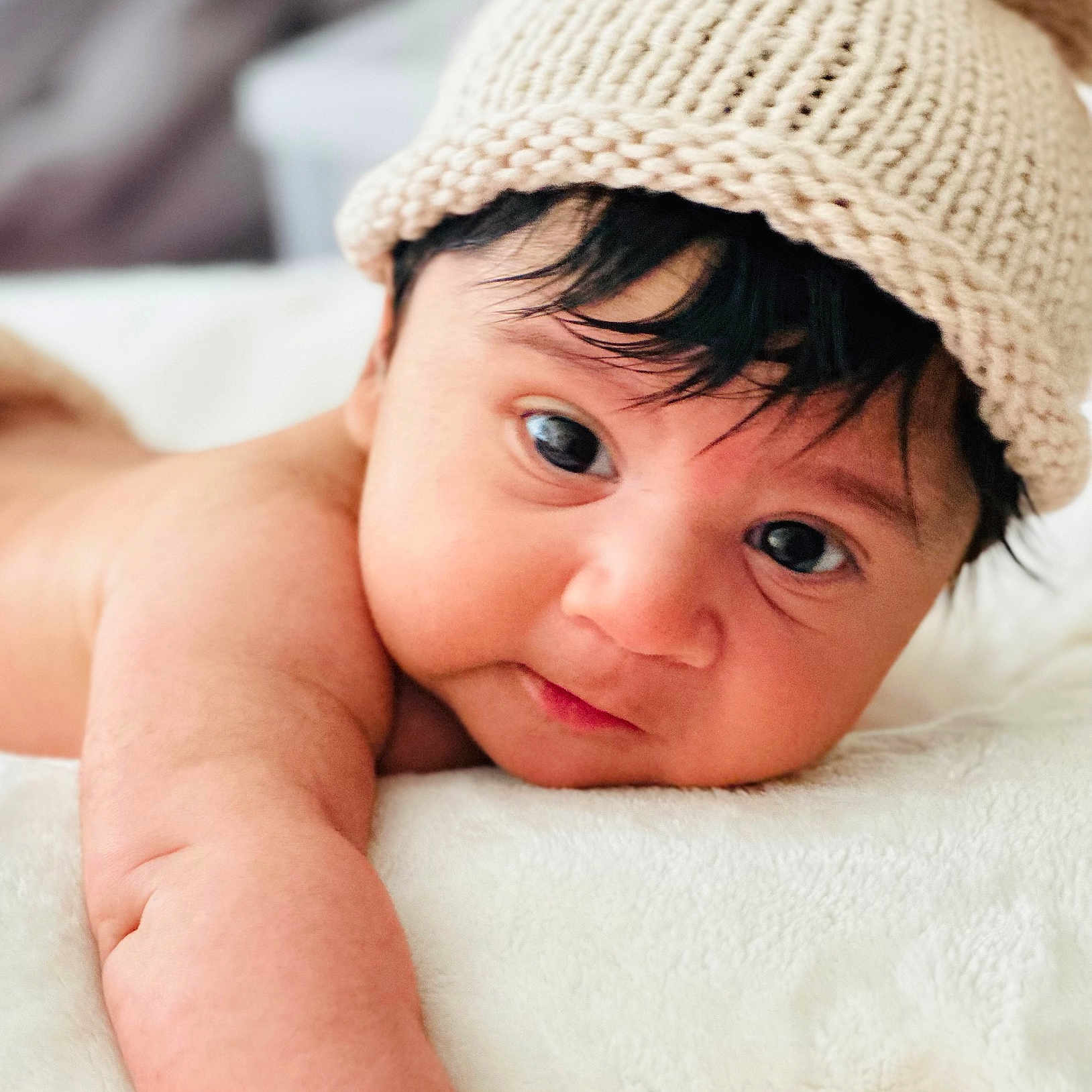 Neville is registered to the contest to win money with this photo: arm, baby, blanket, child, close_up, curious, cute, expression, face, hat, indoors, infant, knitted_hat, lying_down, newborn, person, portrait, skin, sleepwear, soft_texture