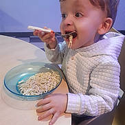 Timéo participe au concours pour gagner de l'argent avec cette photo : child, toddler, eating, noodles, spoon, bowl, table, seat, quilted_jacket, hand, face, mouth_open, happy, meal, indoor, highchair, food, plate, utensil, hair