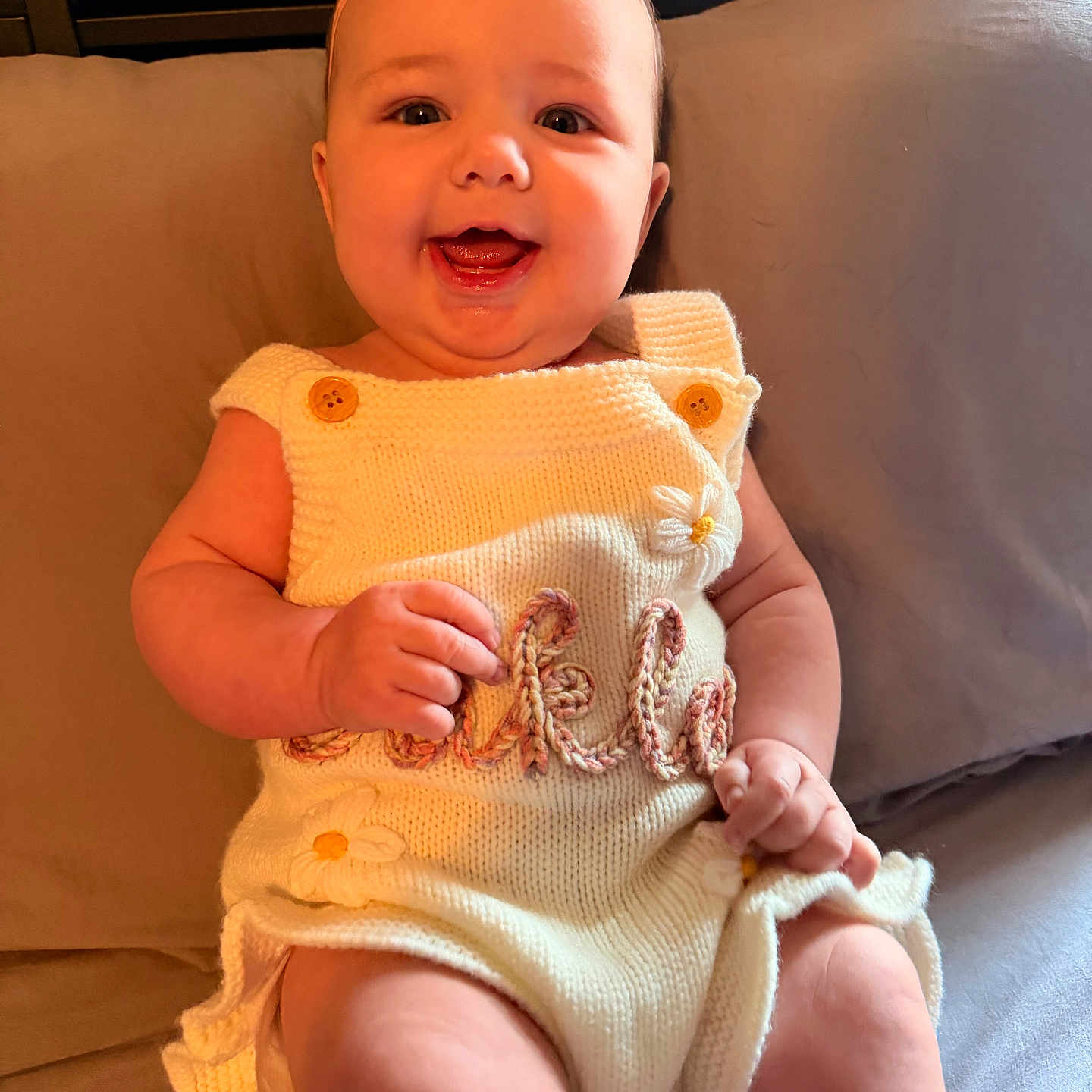 Oakley is registered to the contest to win money with this photo: baby, child, infant, smiling, happy, portrait, sitting, couch, cushion, flower, headband, knitted_clothing, dress, onesie, indoors, warm_lighting, face, hands, legs, photography