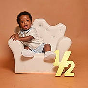 Rushawn joined the competition — help win amazing prizes! toddler, child, armchair, white_chair, plush, half_year, gold_sign, curly_hair, shorts, sneakers, bracelet, neutral_background, studio_photo, portrait, cute, celebration, baby, sitting, expression, indoor