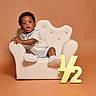 toddler, child, armchair, white_chair, plush, half_year, gold_sign, curly_hair, shorts, sneakers, bracelet, neutral_background, studio_photo, portrait, cute, celebration, baby, sitting, expression, indoor