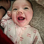 Arya joined the competition — help win amazing prizes! baby, infant, smiling, happy, onesie, patterned_sheet, blanket, bed, face, mouth_open, eyes, eyelashes, rosy_cheeks, hand, adult_hand, wristband, pink, close_up, portrait, sleepwear