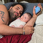 Arya is registered to the contest to win money with this photo: father, baby, infant, bed, pillow, phone, tattoo, bracelet, blanket, cuddling, portrait, indoor, parent_child, family, male, facial_hair, gaze, cozy, skin, hands