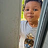 toddler, child, curly_hair, tshirt, bear_graphic, door, peeking, outdoor, porch, sunlight, greenery, smiling, face, person, clothing, playful, young_child, casual_wear, portrait, expression
