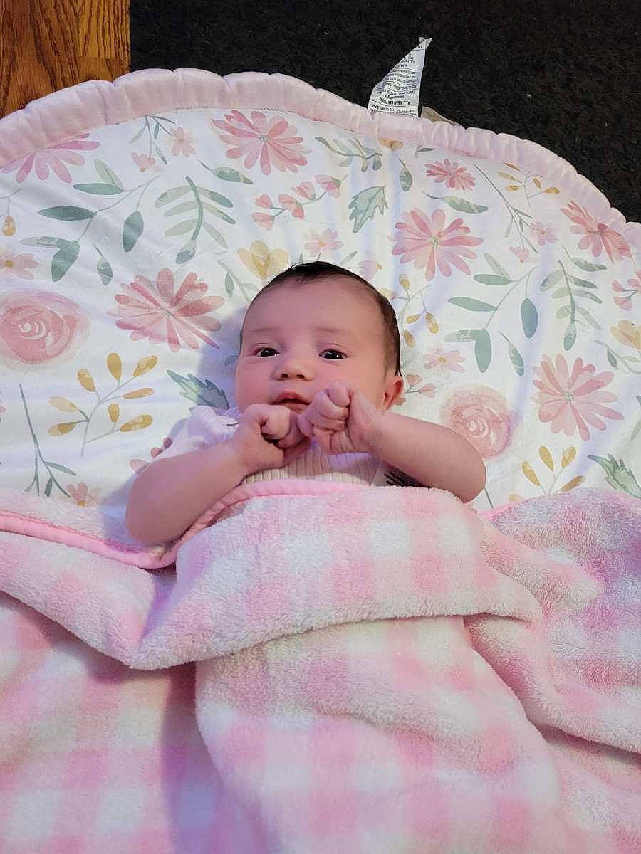 Genevieve is registered to the contest to win money with this photo: baby, baby_products, baby_toddler_clothing, bedding, bedtime, cheek, child, comfort, eye, face, grass, head, linens, pattern, person, pink, room, sitting, skin, textile