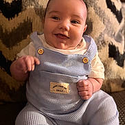 Sebaztian is registered to the contest to win money with this photo: baby, blue_clothing, button, child, closeup, couch, cozy, cute, face, furniture, happy, indoors, infant, overalls, patterned_background, portrait, short_sleeves, sitting, smile, warm_lighting