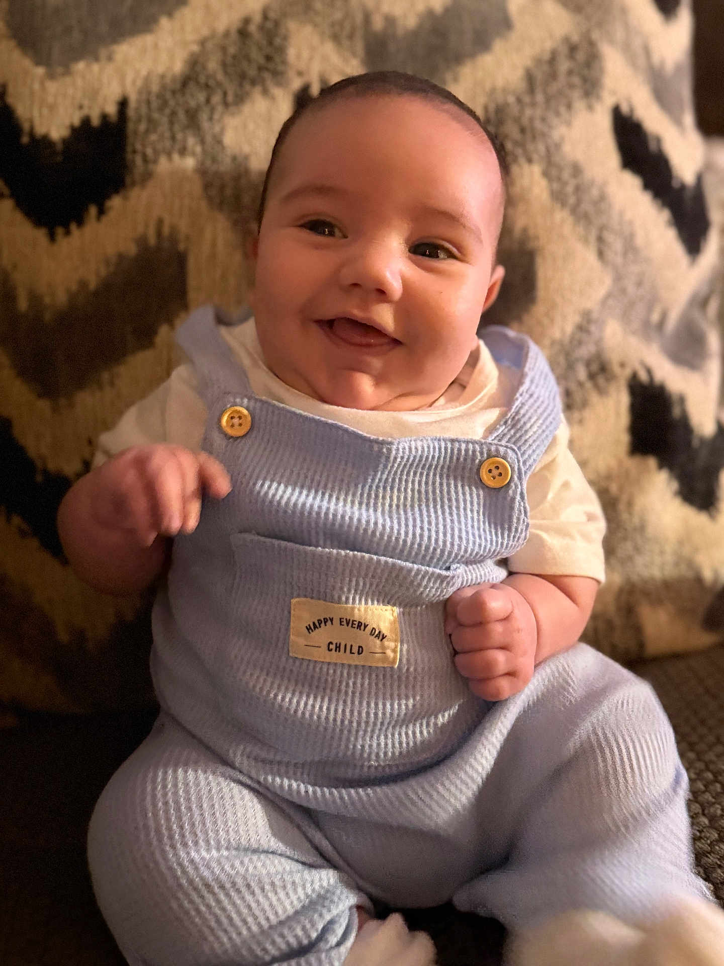 Sebaztian is registered to the contest to win money with this photo: baby, blue_clothing, button, child, closeup, couch, cozy, cute, face, furniture, happy, indoors, infant, overalls, patterned_background, portrait, short_sleeves, sitting, smile, warm_lighting