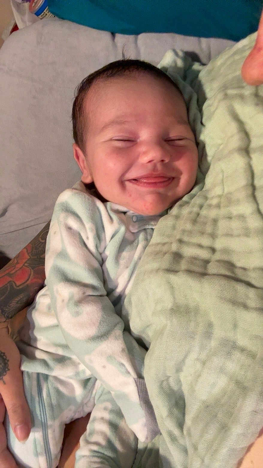 Sebaztian is registered to the contest to win money with this photo: baby, smiling, blanket, tattoo, person, infant, clothing, sleepwear, hand, cuddling, comfort, cute, face, skin, bed, resting, newborn, soft, warm, happy