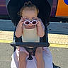 toddler, child, stroller, sunglasses, pink_shoes, yellow_top, outdoor, sunlight, train, platform, sidewalk, playful, person, transportation, summer, casual_clothing, cute, baby, seat, urban