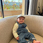 Dominique participe au concours pour gagner de l'argent avec cette photo : baby, denim_overalls, cap, armchair, indoors, window, plants, balcony, sunlight, cozy, smiling, child, barefoot, curtains, striped_shirt, furniture, relaxing, portrait, casual_clothing, home