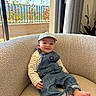 baby, denim_overalls, cap, armchair, indoors, window, plants, balcony, sunlight, cozy, smiling, child, barefoot, curtains, striped_shirt, furniture, relaxing, portrait, casual_clothing, home