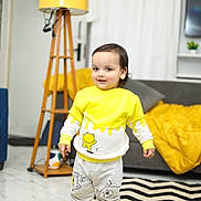 Kriyansh is registered to the contest to win money with this photo: toddler, child, yellow_clothing, sneakers, cushion, lamp, living_room, sofa, blanket, rug, cartoon_design, floor, curtains, toy_keyboard, smiling, indoor, person, home_decor, wooden_lamp_stand, happy