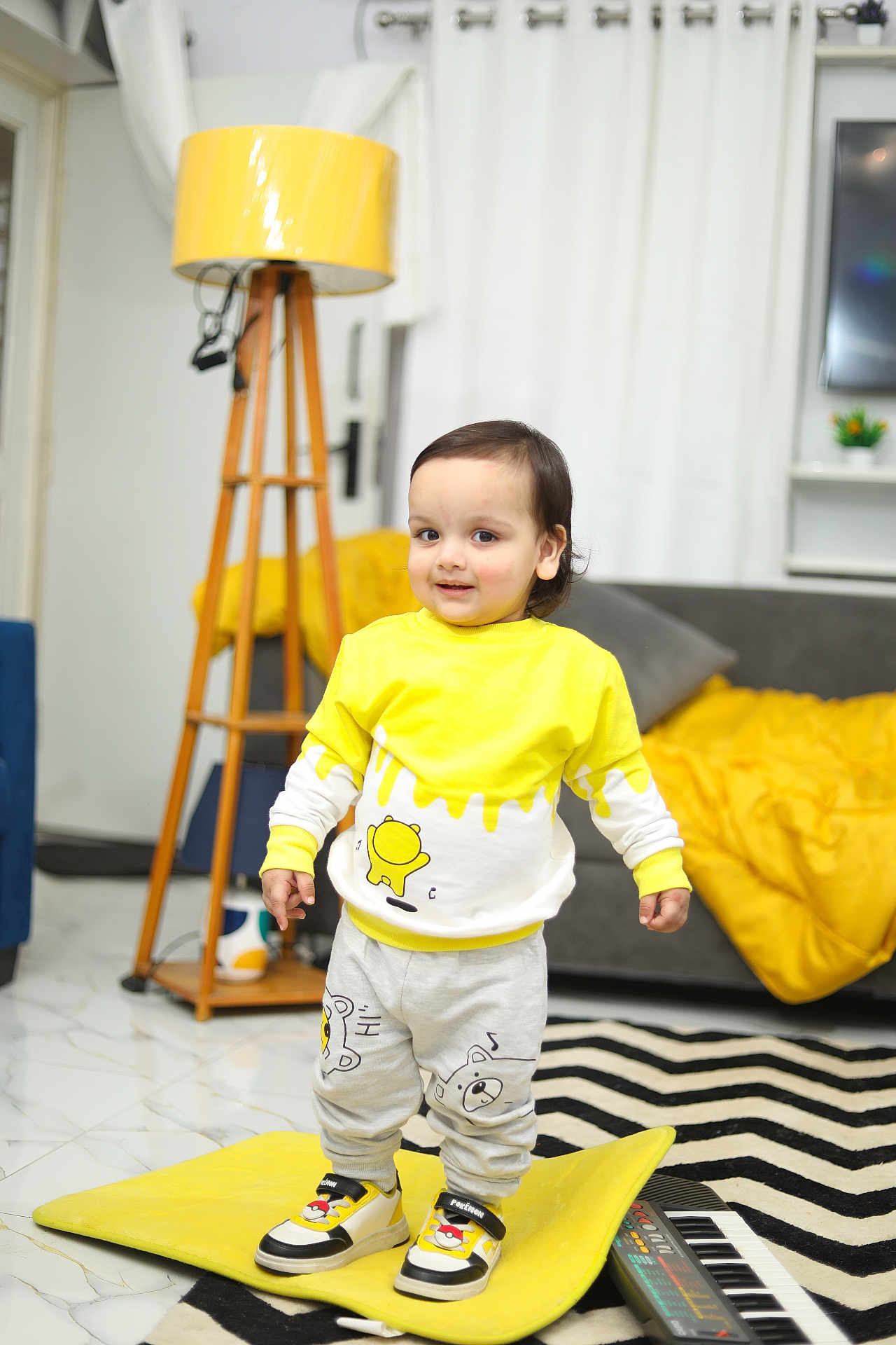 Kriyansh is registered to the contest to win money with this photo: toddler, child, yellow_clothing, sneakers, cushion, lamp, living_room, sofa, blanket, rug, cartoon_design, floor, curtains, toy_keyboard, smiling, indoor, person, home_decor, wooden_lamp_stand, happy