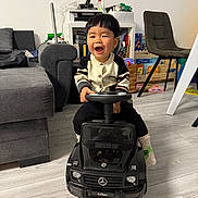 Isaac a rejoint le concours — aidez-le/la à gagner de superbes lots ! child, toddler, toy_car, indoor, living_room, smiling, happy, black_toy, mercedes, g_class, sweater, socks, flooring, sofa, chair, clock, cabinet, books, playroom, furniture