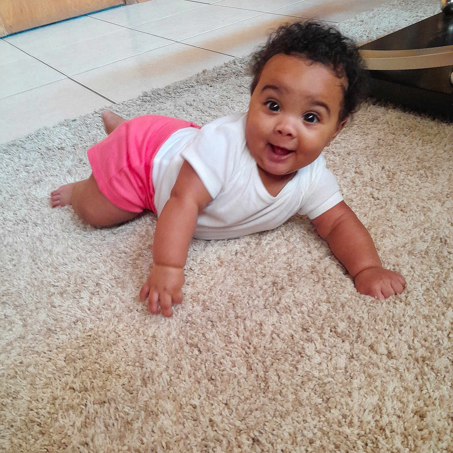 Ni'Amani joined the competition — help win amazing prizes! baby, carpet, child, crawling, curly_hair, cute, eyes, face, floor, hand, happy, indoor, infant, person, pink_shorts, skin, smile, tile_floor, white_shirt, wooden_door