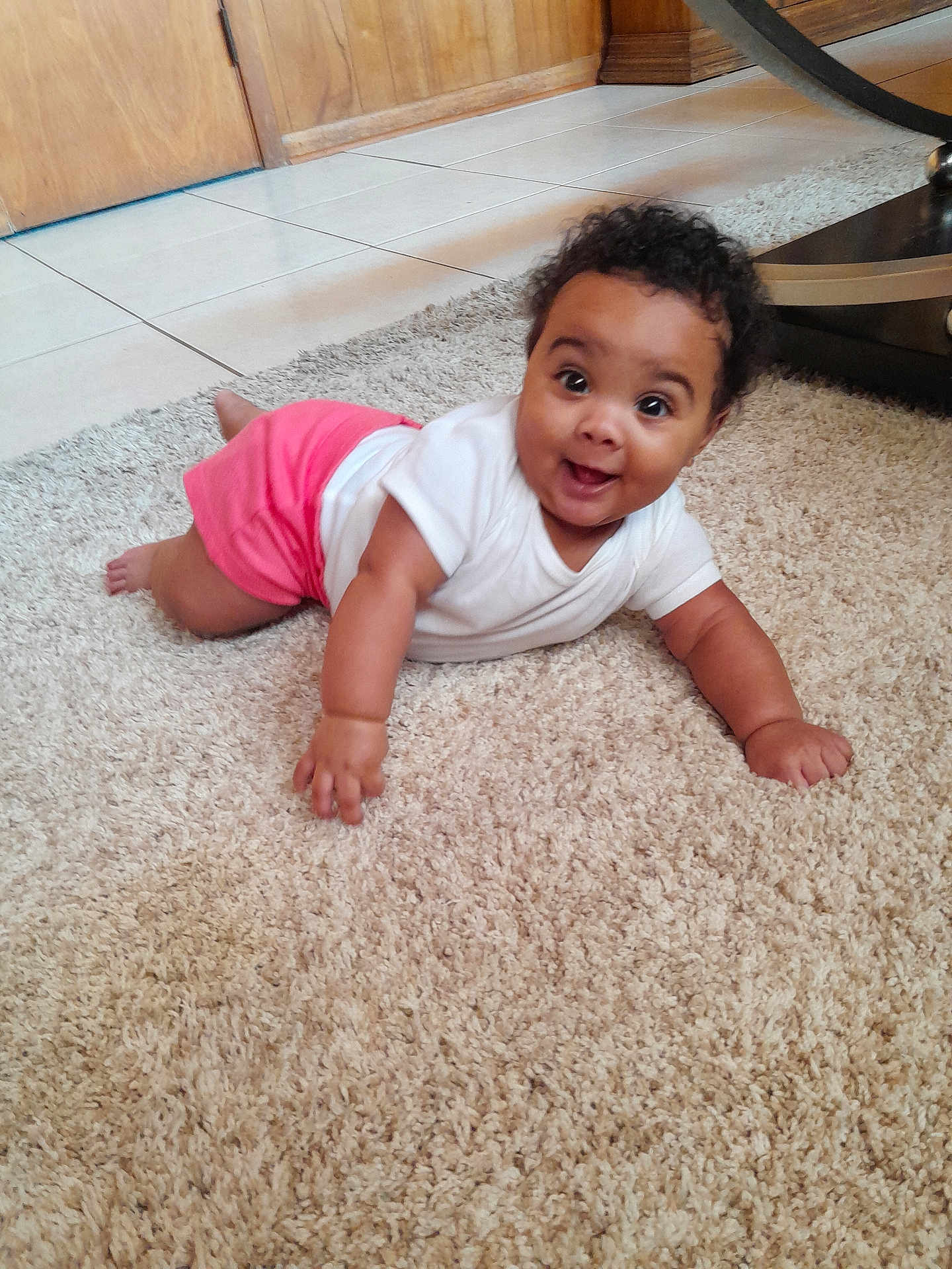 Ni'Amani joined the competition — help win amazing prizes! baby, carpet, child, crawling, curly_hair, cute, eyes, face, floor, hand, happy, indoor, infant, person, pink_shorts, skin, smile, tile_floor, white_shirt, wooden_door