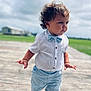 baby, blue_shorts, bow_tie, child, cloudy_sky, curly_hair, cute, daylight, fashion, green_field, outdoor, person, portrait, serious_expression, standing, summer_clothing, toddler, walking, white_shirt, wooden_floor