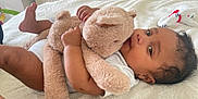 Dylan is registered to the contest to win money with this photo: baby, teddy_bear, blanket, indoor, child, soft_toy, lying_down, curly_hair, white_clothing, plush, comfort, cozy, cute, infant, bedroom, person, sleepwear, toy, resting, soft_texture
